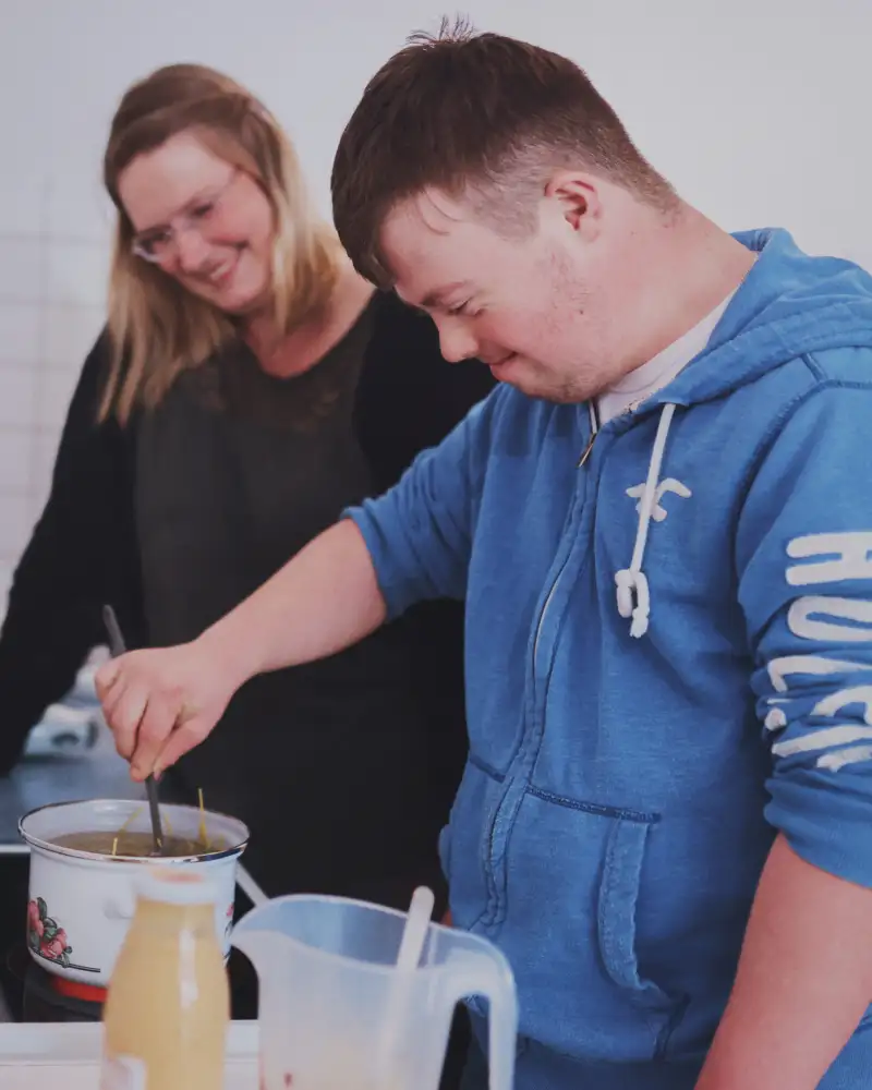 Ein junger Mann mit Downsyndrom rührt in einem Suppentopf. Neben ihm steht eine Frau und schaut zu.