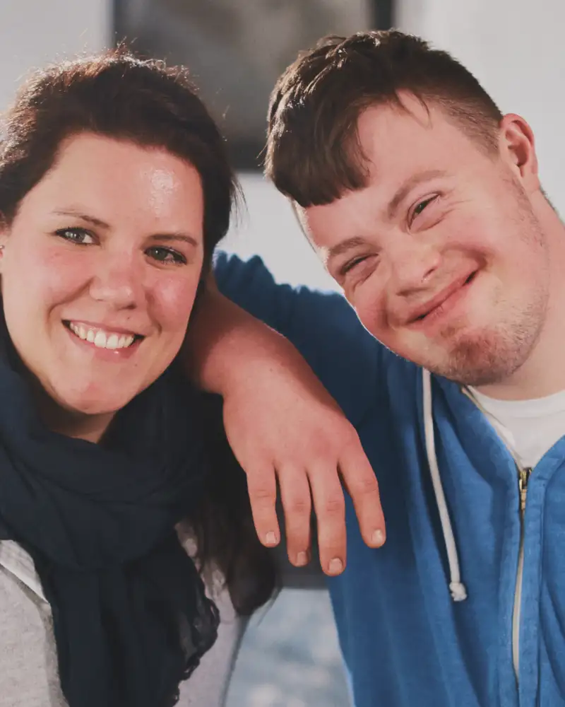 Ein Mann mit Downsyndrom und eine Frau lächeln in die Kamera.