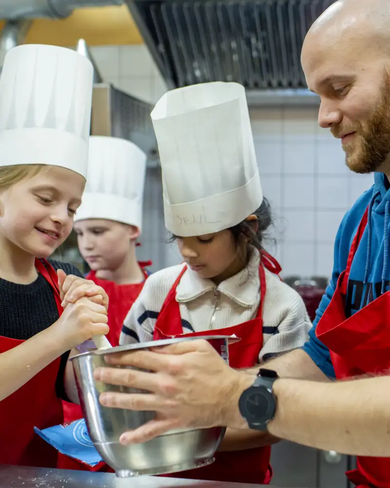 Ein Mann mit Schürze und drei Kinder mit Schürzen und Kochmützen in einer Küche