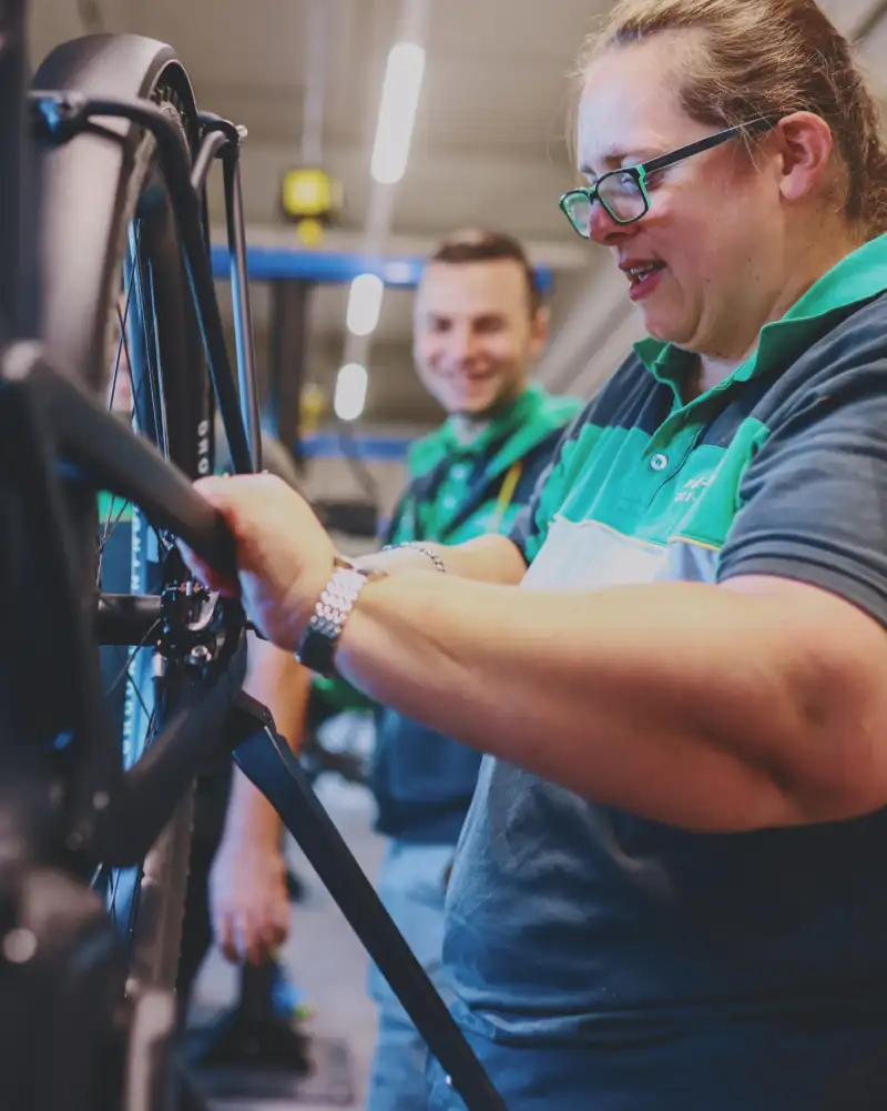 Eine Frau in Arbeitskleidung repariert ein aufgebocktes E-Bike. Im Hintergrund steht ein Mann.