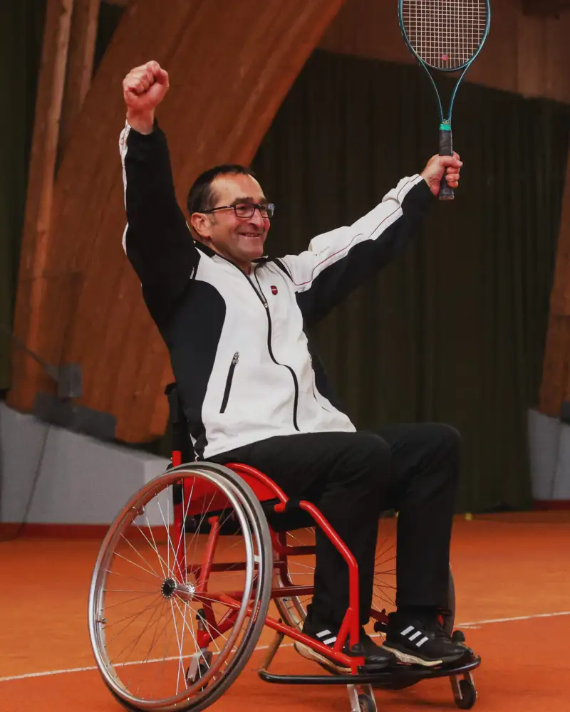 Ein Mann im Sportrollstuhl reißt beide Arme hoch und lacht. In der einen Hand hält er einen Tennisschläger.