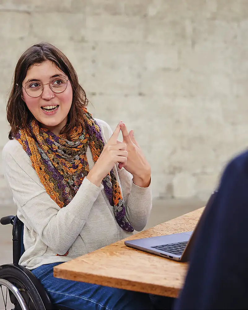 Eine Frau im Rollstuhl sitzt hinter einem Schreibtisch. Sie spricht mit einer Person, die vor dem Schreibtisch sitzt.