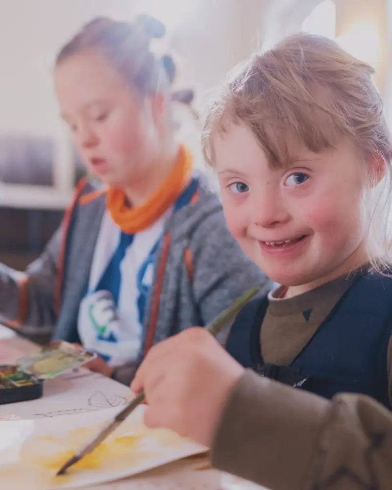 Zwei Mädchen mit Downsyndrom sitzen nebeneinander am Tisch und malen mit Wasserfarbe auf Papier.