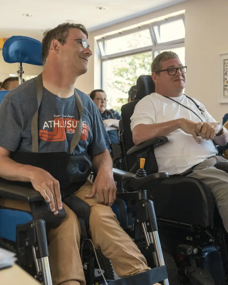Personen unterschiedlichen Alters sitzen in einem Vortragsraum. Im Vordergrund sind zwei Männer im Rollstuhl.