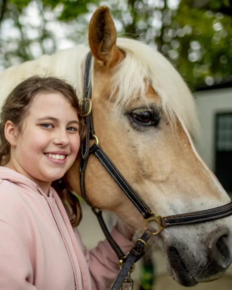 Ein Mädchen schmiegt ihren Kopf an den Kopf eines Ponys. Dabei hält sie sein Zaumzeug in der Hand.