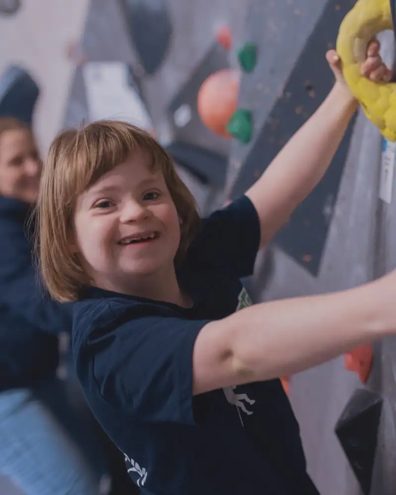 Ein Mädchen mit Downsyndrom klettert lächelnd an einer Boulderwand