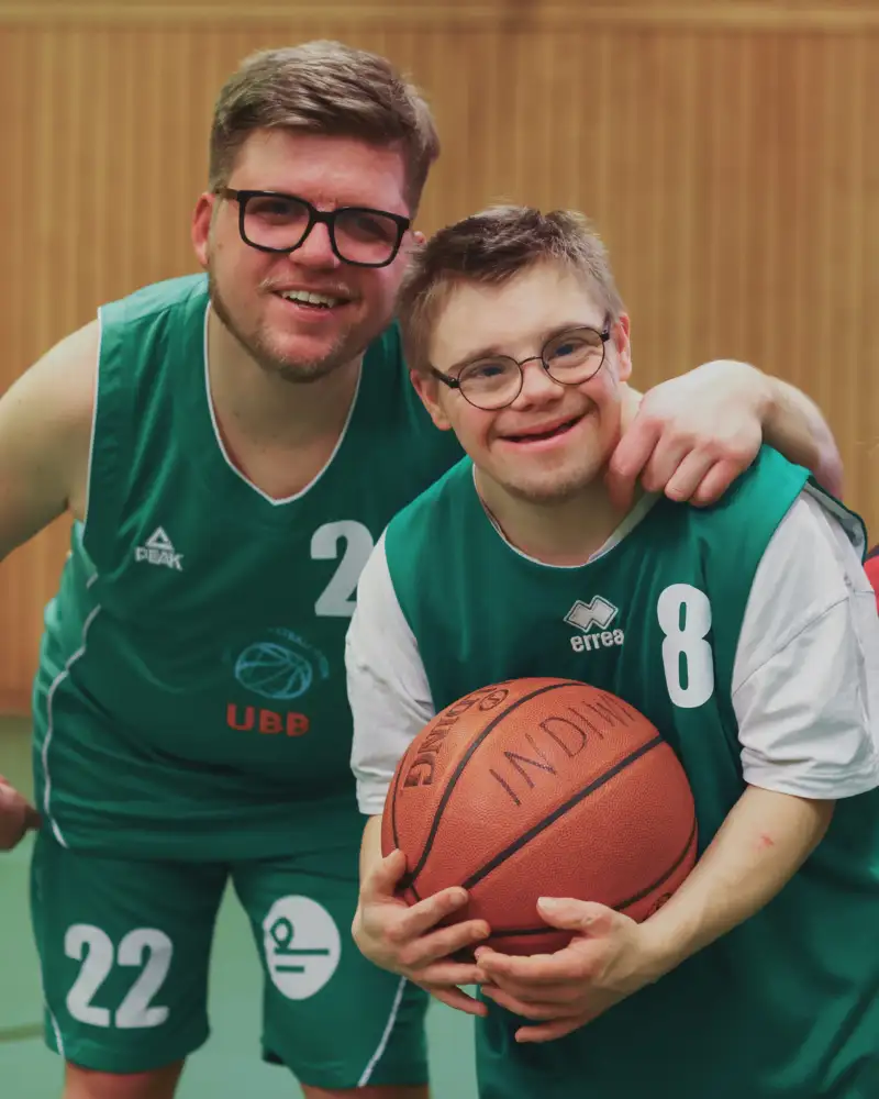 Drei Personen im Sportdress. Einer von ihnen hat das Downsyndrom. Er hält einen Basketball in den Händen.