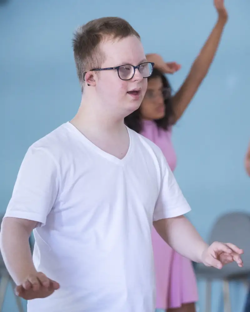 Ein junger Mann mit Downsyndrom in einem Tanzsaal. Neben ihm stehen zwei Frauen. Die drei bewegen sich offenbar zu einer Musik.