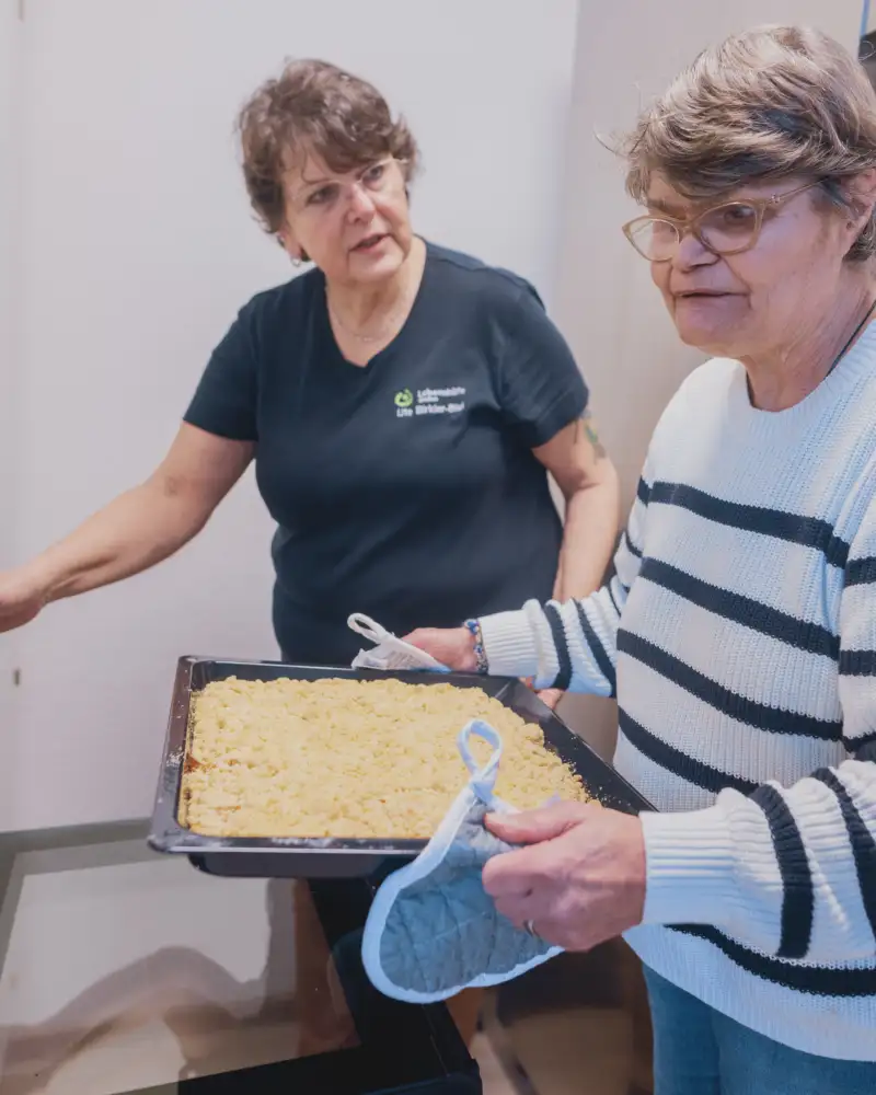 Eine ältere Frau trägt ein Blech mit Streuselkuchen zum Backofen. Neben ihr steht eine Frau im mittleren Alter.