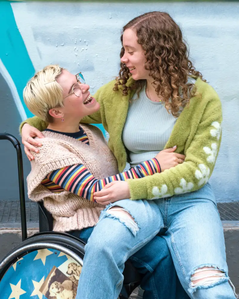 Ein Mädchen sitzt auf dem Schoß eines anderen Mädchens, das im Rollstuhl sitzt. Die beiden umarmen sich.