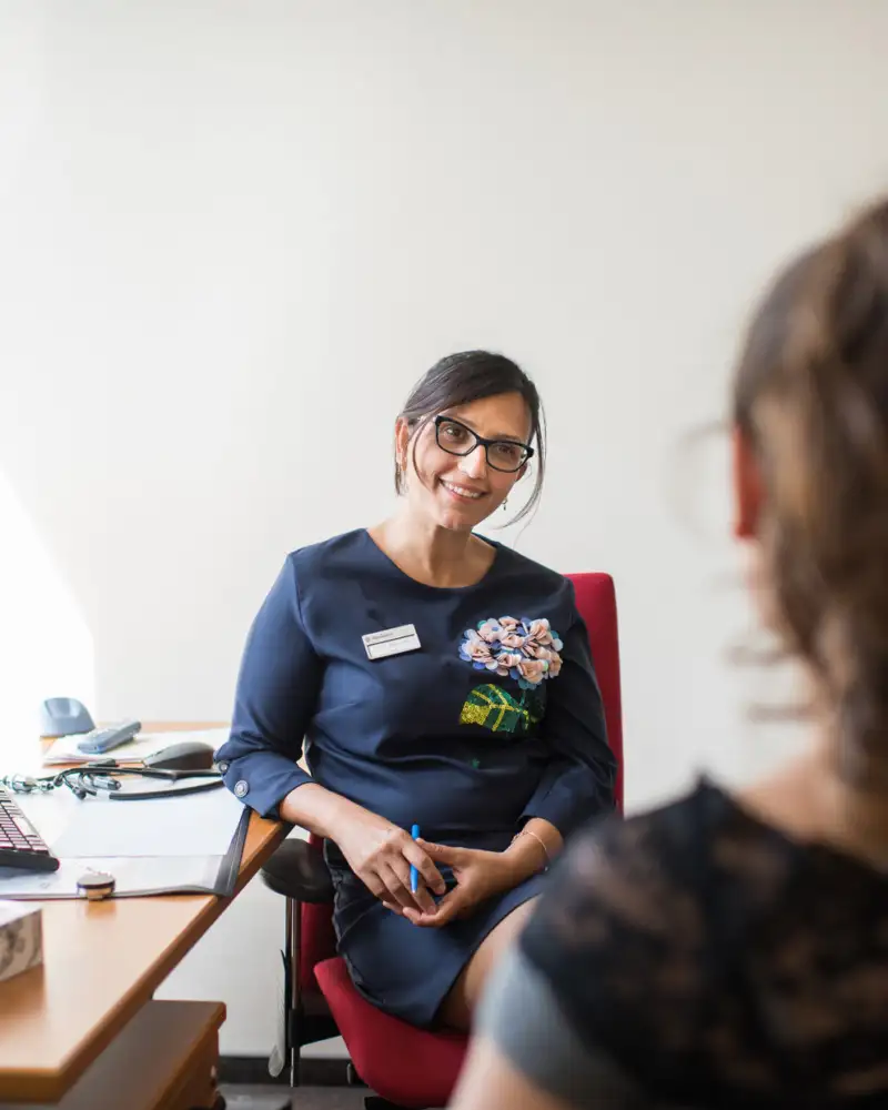 Eine junge Frau mit Brille sitzt am Schreibtisch, ihr gegenüber sitzt eine andere Person, die nur von hinten zu sehen ist.
