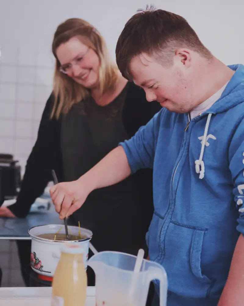 Ein junger Mann mit Downsyndrom steht in einer Küche und rührt in einem Topf. Eine Frau schaut ihm dabei zu.