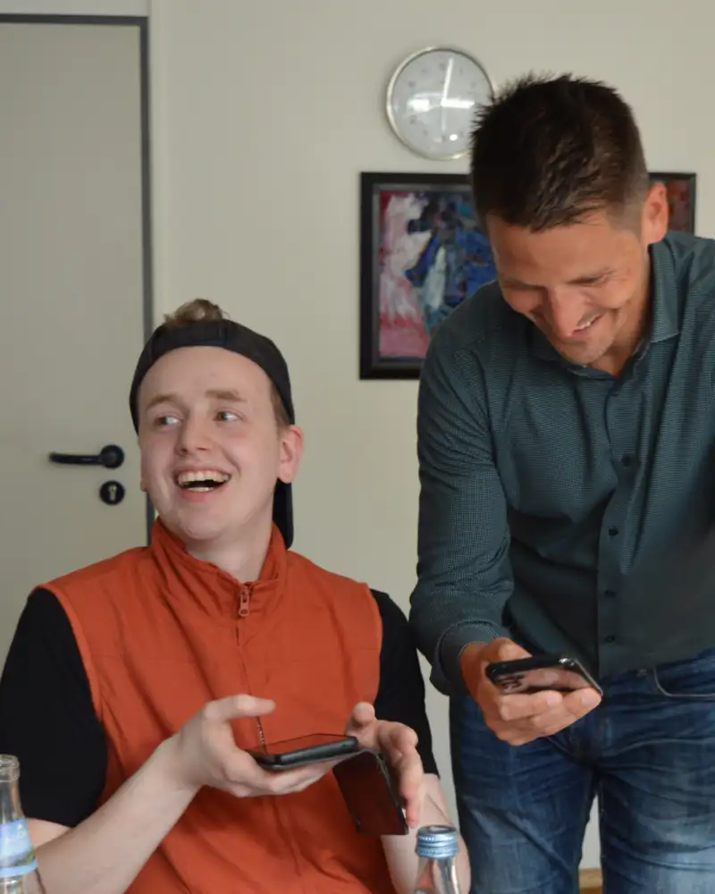 In einem Zimmer an einem Esstisch sind zwei Männer, die beide ein Handy in der Hand halten. Der jüngere von beiden ist sportlich gekleidet mit falsch herum aufgesetzter Kappe. Er schaut auf und lacht. Der altere in Jeans und Hemd steht neben ihm, einen Arm auf den Tisch gestützt und schaut auf sein Handy.