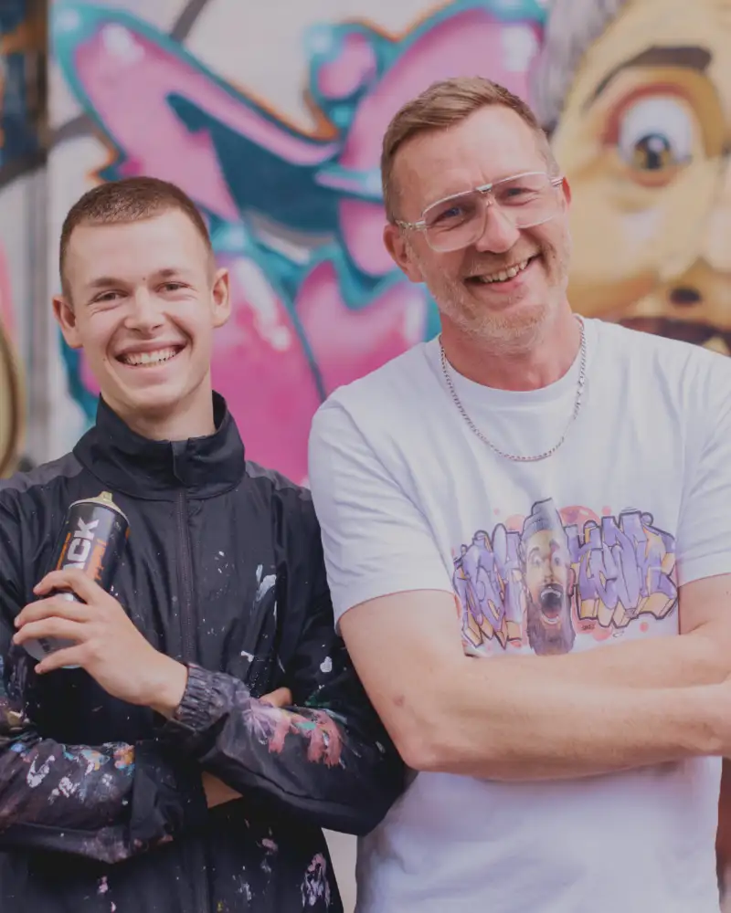 Ein Jugendlicher mit Spraydose und ein Mann stehen lächelnd vor einer Graffiti-Wand