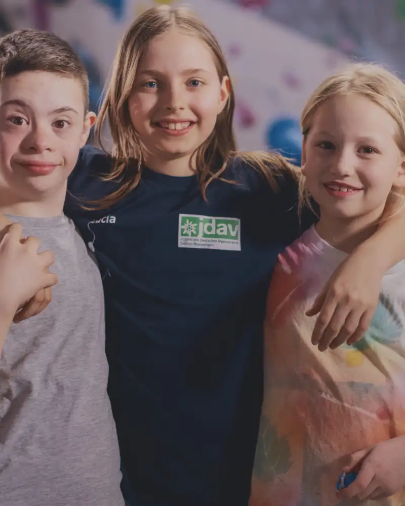 Zwei Mädchen und ein Junge mit Downsyndrom. Das Mädchen in der Mitte hat ihre Arme um die Schultern der anderen gelegt.