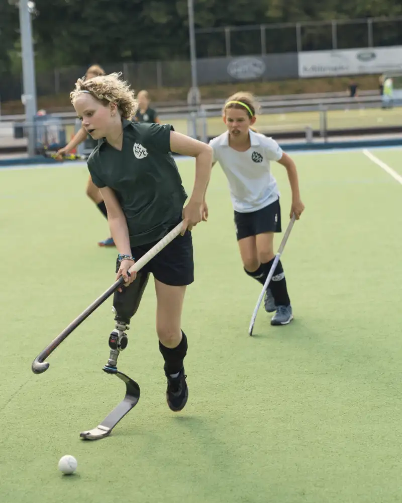Zwei junge Mädchen, eine von ihnen mit Beinprothese, spielen zusammen Hockey auf einem Sportplatz im Freien.