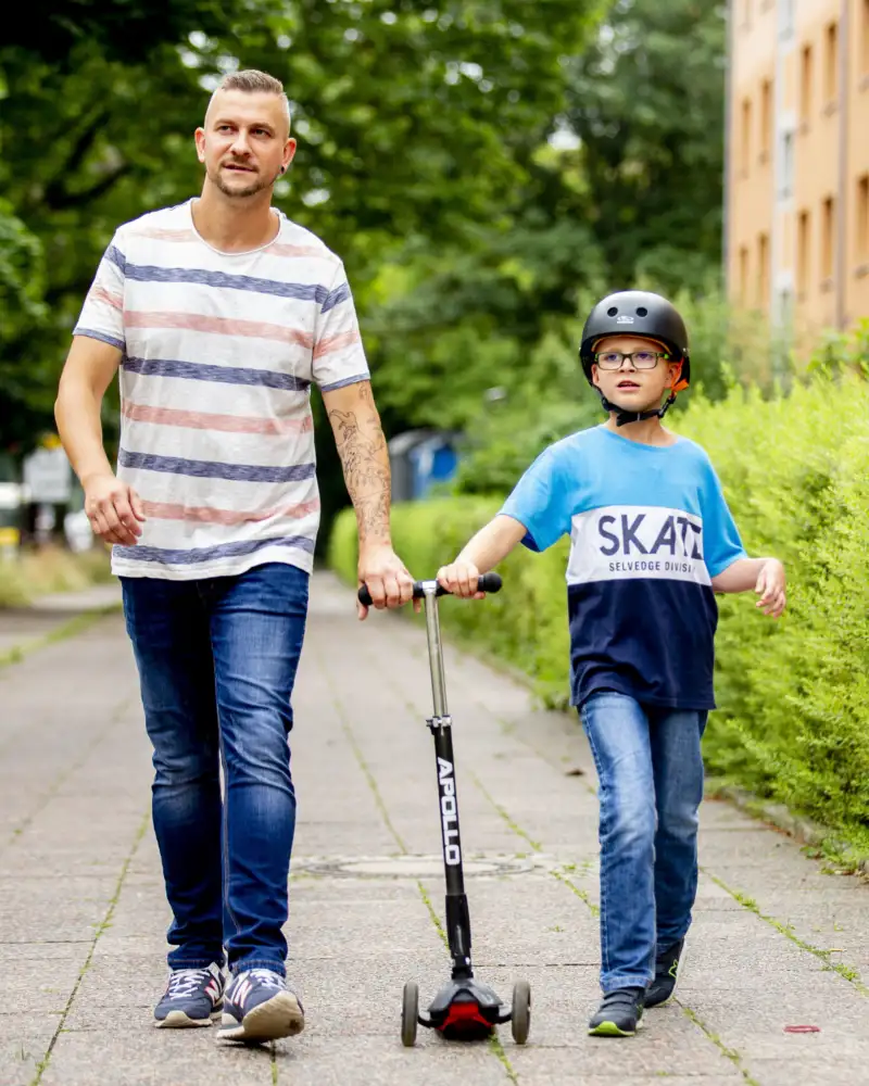 Ein Mann mit Jeans und T-Shirt und ein Junge sind zusammen in der Stadt unterwegs. Der Junge trägt einen Helm. Zusammen schieben sie einen Roller.
