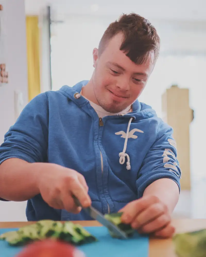 Ein junger Mann mit Downsyndrom im blauen Hoodie und modischer Kurzhaarfrisur schnippelt in einer Küche Zucchini.