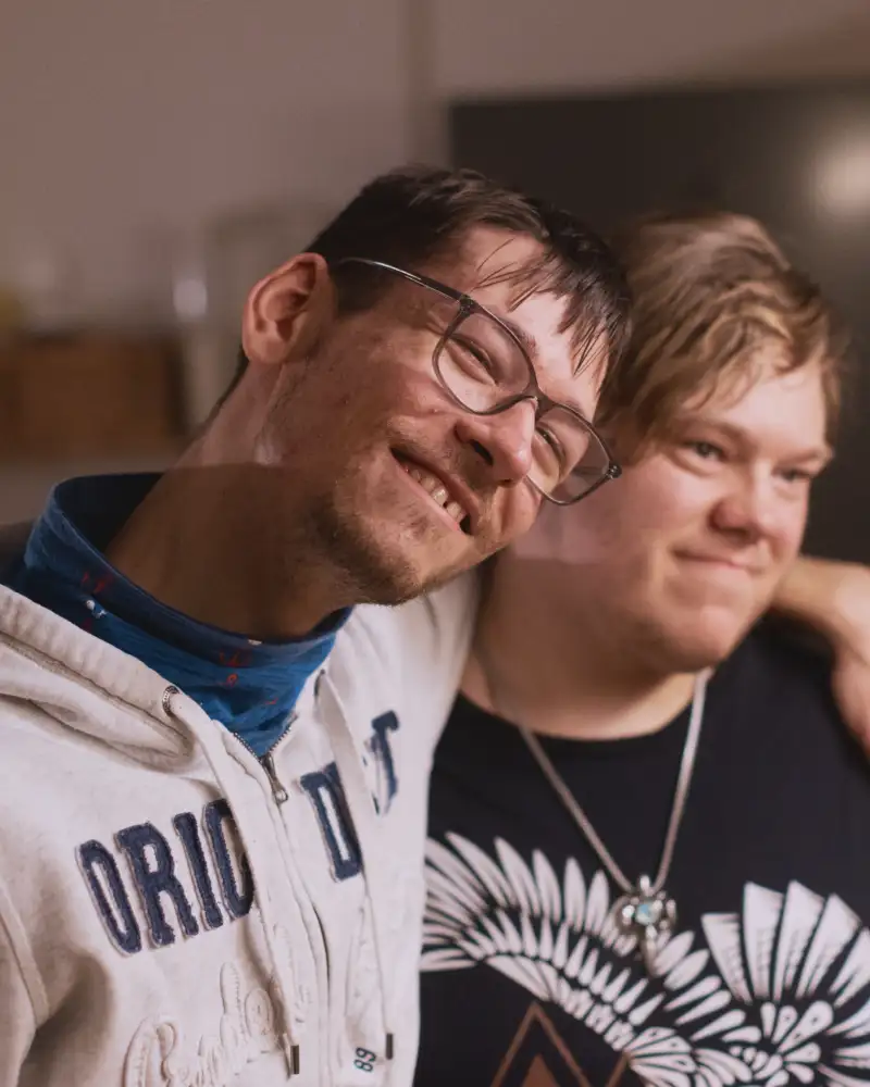 Zwei junge Männer mit geistiger Behinderung stehen lächelnd Arm in Arm nebeneinander.
