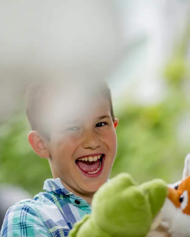 Ein Junge hält zwei Handpuppen hoch, die verschiedene Tiere darstellen. Er lacht in die Kamera. Hinter ihm steht ein Jugendlicher.