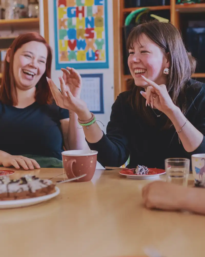 Drei junge Frauen sitzen bei Kaffee und Kuchen an einem Tisch und lachen miteinander.