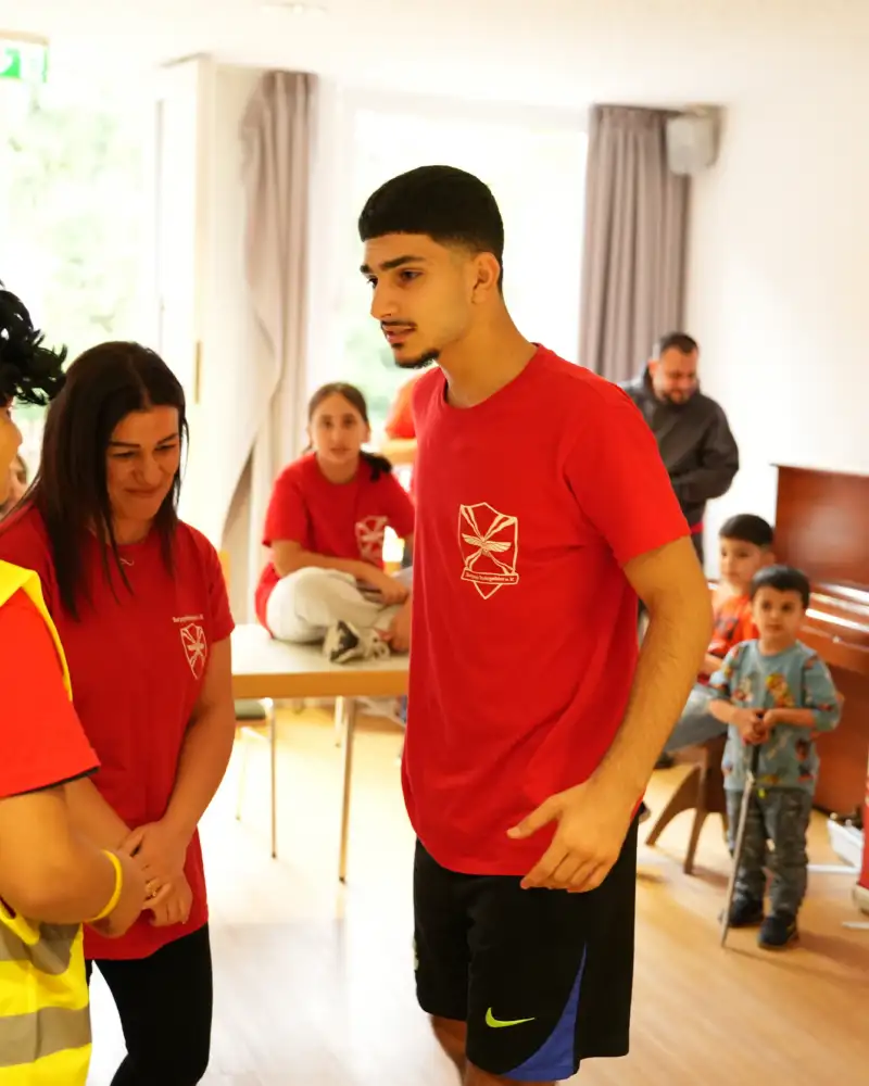 Theaterprobe: Drei Jugendliche stehen in einem Raum beieinander. Einer von ihnen ist im Begriff etwas zu sagen. Im Hintergrund schauen einige Kinder zu.