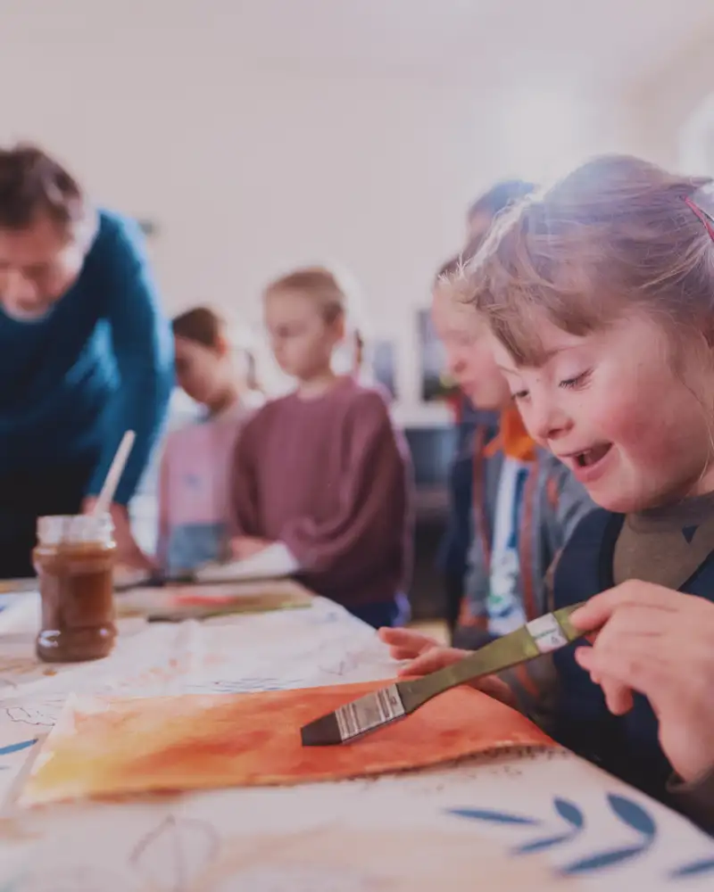 Ein Mädchen mit Downsyndrom mal mit Aquarellfarbe. Im Hintergrund sind weitere Kinder zu sehen, die von einem Erwachsenen beim Malen angeleitet werden.