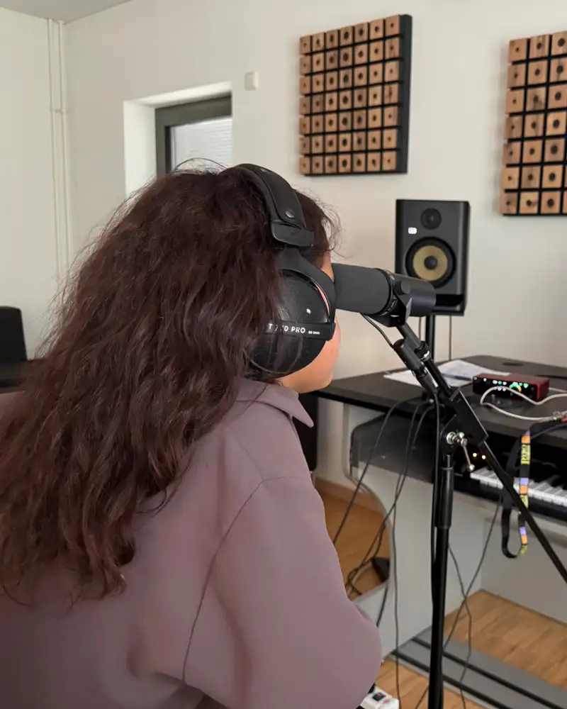 Eine Jugendliche sitzt mit Kopfhörern in einem Studio vor einem Mikrofon. Ihr gegenüber steht ein Monitor, auf dem unscharf Skalen und Tabellen gezeigt werden. Die Jugendliche ist nur von hinten zu sehen.