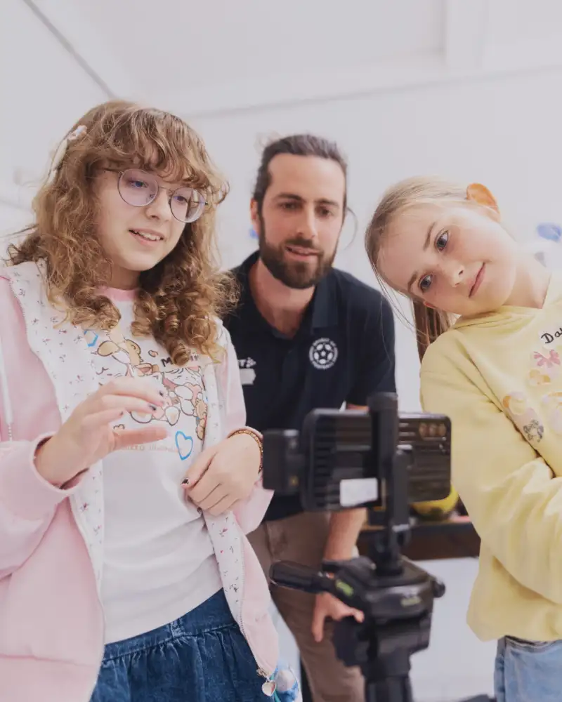 Zwei Mädchen drehen einen Film mit einem Handy, im Hintergrund schaut ein junger Mann zu