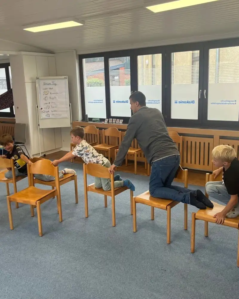 Drei kleine Jungen und ein Mann krabbeln nacheinander über fünf Stühle. Ein sechster Stuhl ist leer. Mehr Jungen warten hinter der Gruppe, bis sie dran sind. Eine Frau schaut zu.