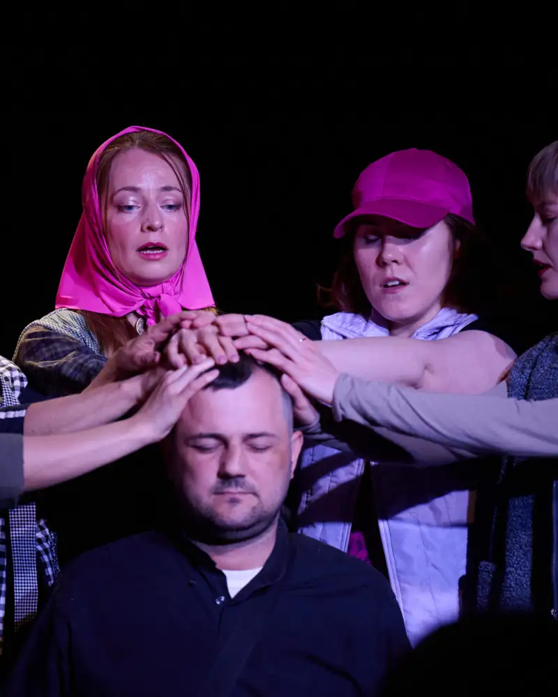 Theaterszene: 5 Frauen mit pinkem Kopftuch legen die Hand auf den Kopf eines Mannes