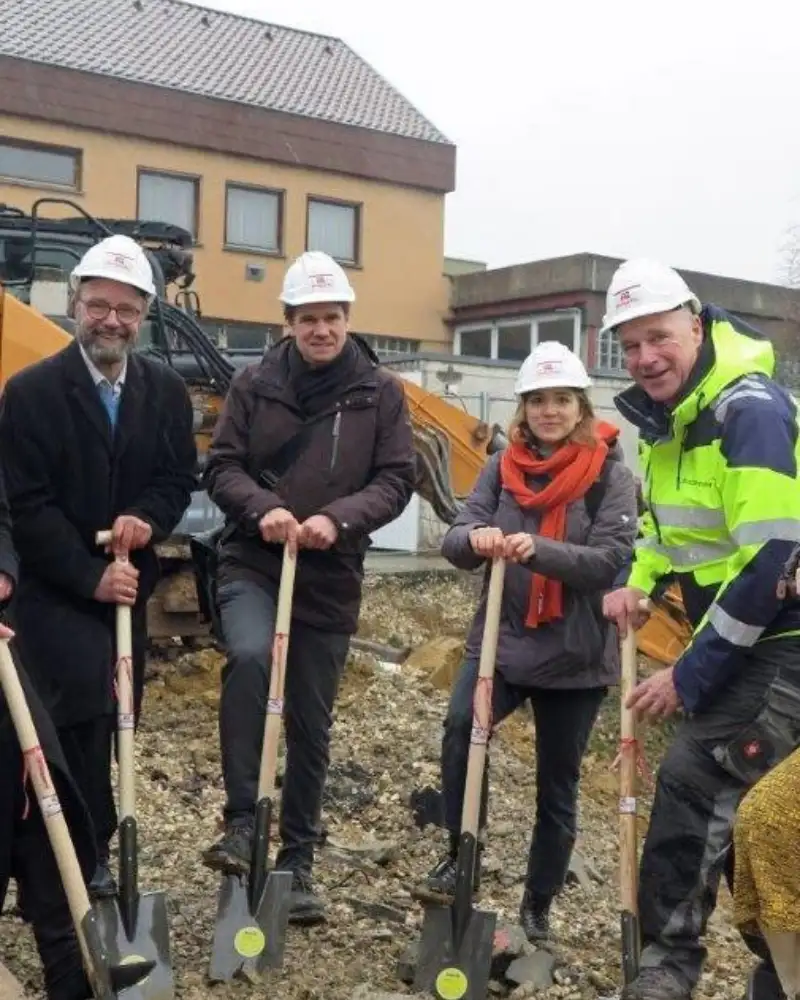 In einem Halbkreis stehen drei Frauen und drei Männer auf einer Baustelle. Alle tragen Schutzhelme und stützen sich auf Spaten.