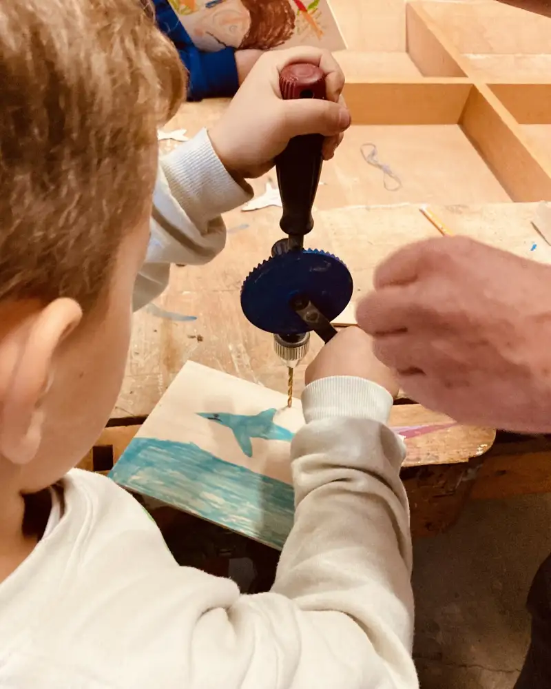 Ein Kleiner Junge bohrt mit einem Handbohrer ein Loch in ein kleines Bild mit einem Haifisch. Neben ihm steht eine erwachsene Person. Die beiden stehen an einem Arbeitstisch.