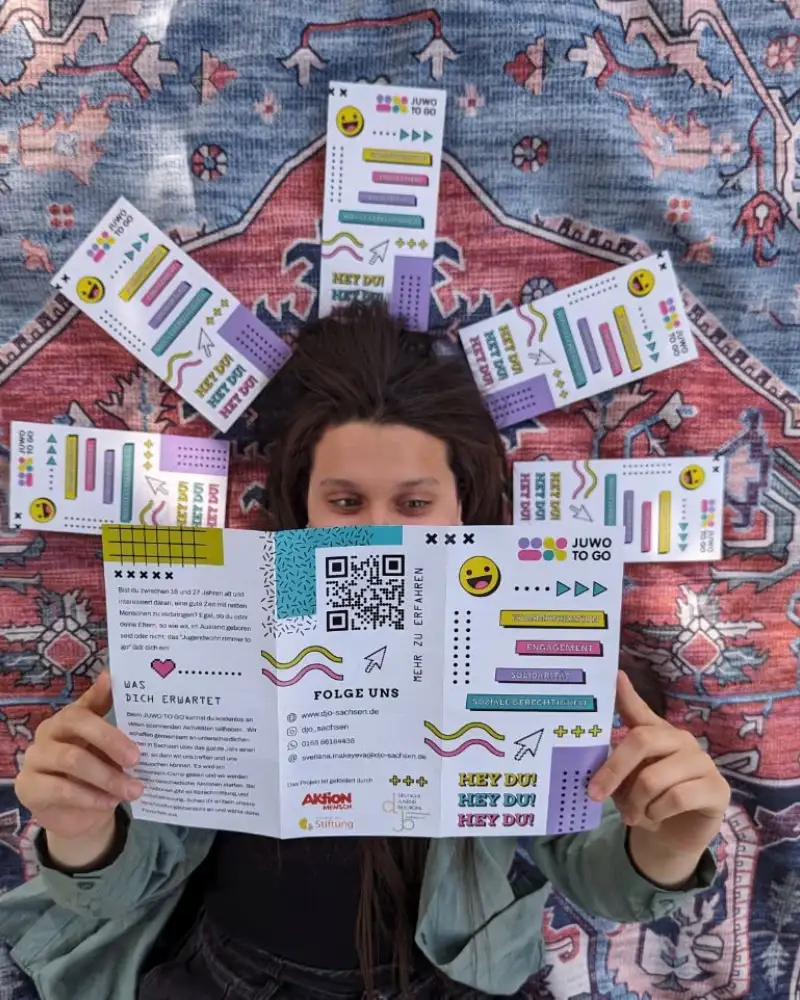 Eine junge Frau liegt auf dem Rücken auf einem bunten Teppich. Um ihren Kopf herum liegen fünf zusammengeklappte Flyer. Den sechsten Flyer liest sie. Man erkennt darauf unter anderem die Aufschrift JUWO TO GO. Sie hat die drei Seiten des Flyers aufgeklappt und hält ihn über sich, so dass er die untere Hälfte ihres Gesichts verdeckt.