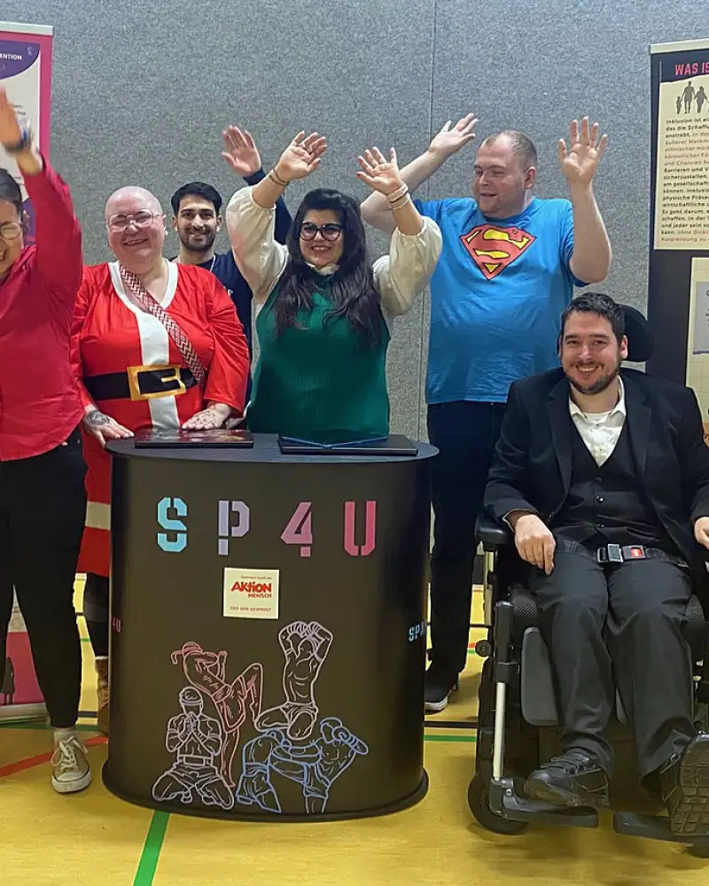 Eine jubelnde Gruppe Menschen mit und ohne Behinderung hinter einem Stand in einer Sporthalle.
