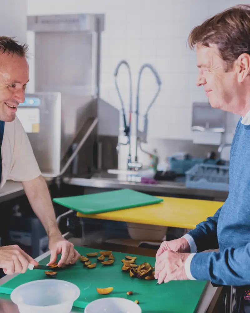 Rudi Cerne und ein Mann schneiden gemeinsam saure Gurken klein auf einem grünen Schneidbrett.