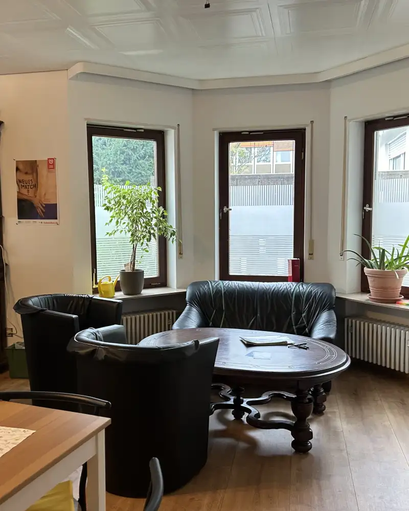 In einem Raum steht eine Sitzgruppe in einem Erker mit drei Fenstern. Links davon steht ein großer Buffetschrank aus dunklem Holz im Gründerzeitstil.