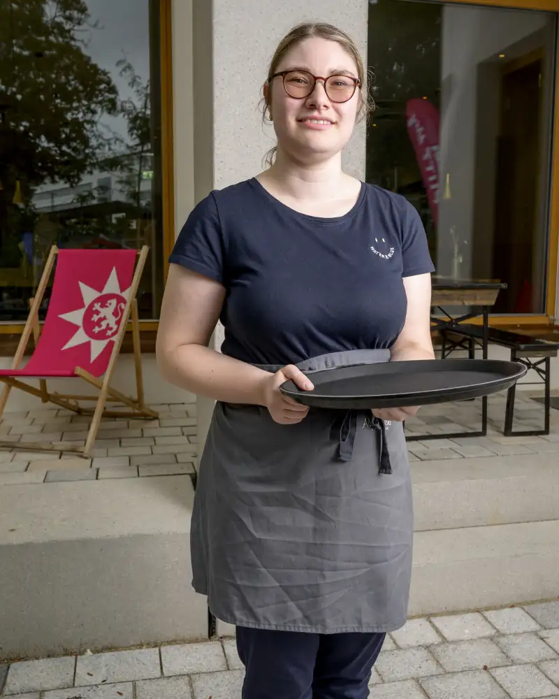 Eine junge Frau mit getönter Brille steht auf einer Terrasse vor einem Gebäude. Sie trägt eine Schürze und hält ein Tablett in den Händen..