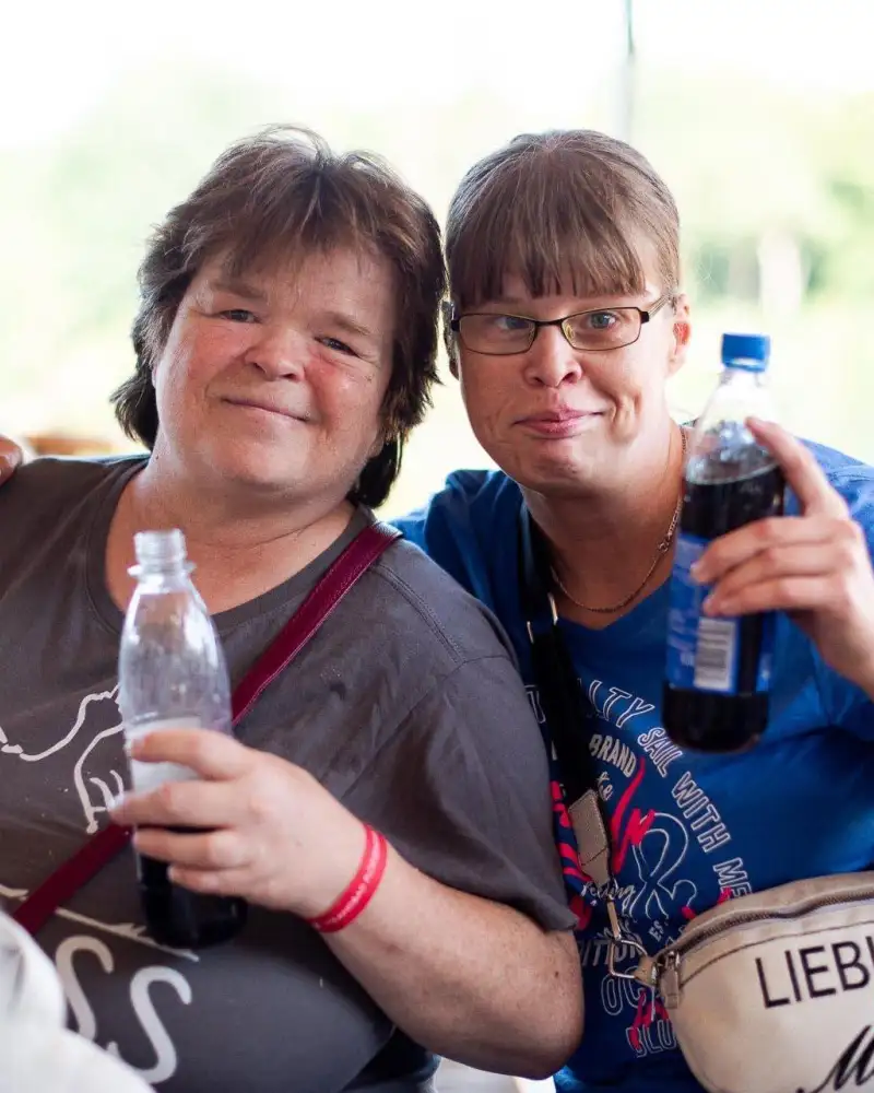 Eine Frau legt den Arm um die Schultern einer anderen. Beide halten Softdrinks in der Hand.