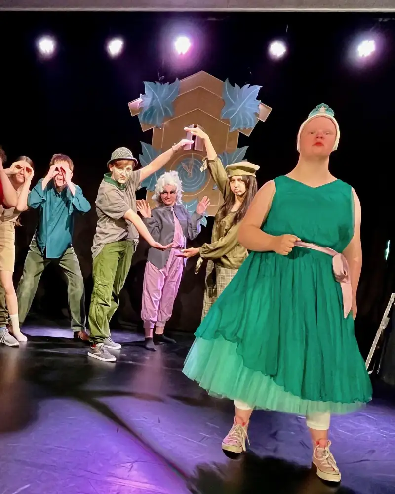 Auf einer Theaterbühne stehen acht Jugendliche in Kostümen. Zwei von ihnen stehen im Vordergrund. Die eine ist mit einer Trägerhose, Bluse und Weste bekleidet.  Sie hat ein Buch unterm Arm. Die andere, eine junge Frau mit Downsyndrom, trägt ein Ballkleid.
