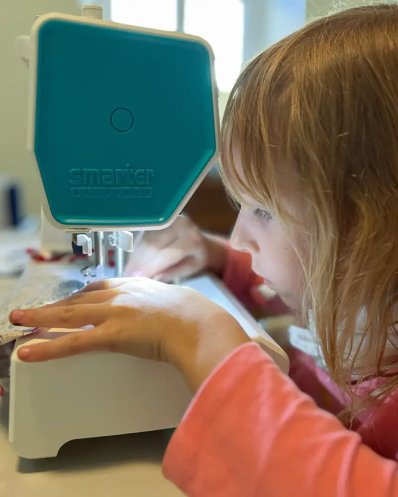 Ein kleines Mädchen mit langen Haaren ist sehr konzentriert bei der Arbeit an einer Nähmaschine.