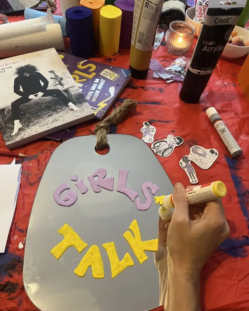Auf einem Tisch liegen Bücher, farbige Rollen aus Krepppapier, Klebstofftuben, ausgeschnittene Zeichnungen und ein Spiegel. Auf dem Spiegel sind ausgeschnittene Buchstaben aus buntem Filz aufgeklebt. Sie bilden die Worte Girls Talk.