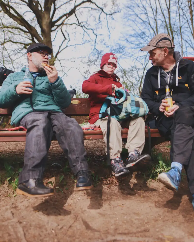 Zwei Männer und eine Frau mit Down Syndrom sitzen zusammen auf einer Bank im Wald und essen Snacks.