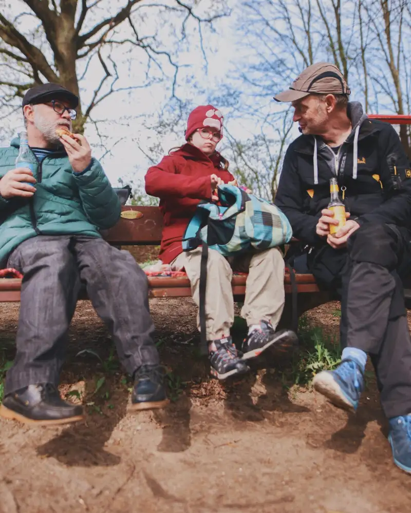 Zwei Männer und eine Frau mit Down Syndrom sitzen zusammen auf einer Bank im Wald und essen Snacks.