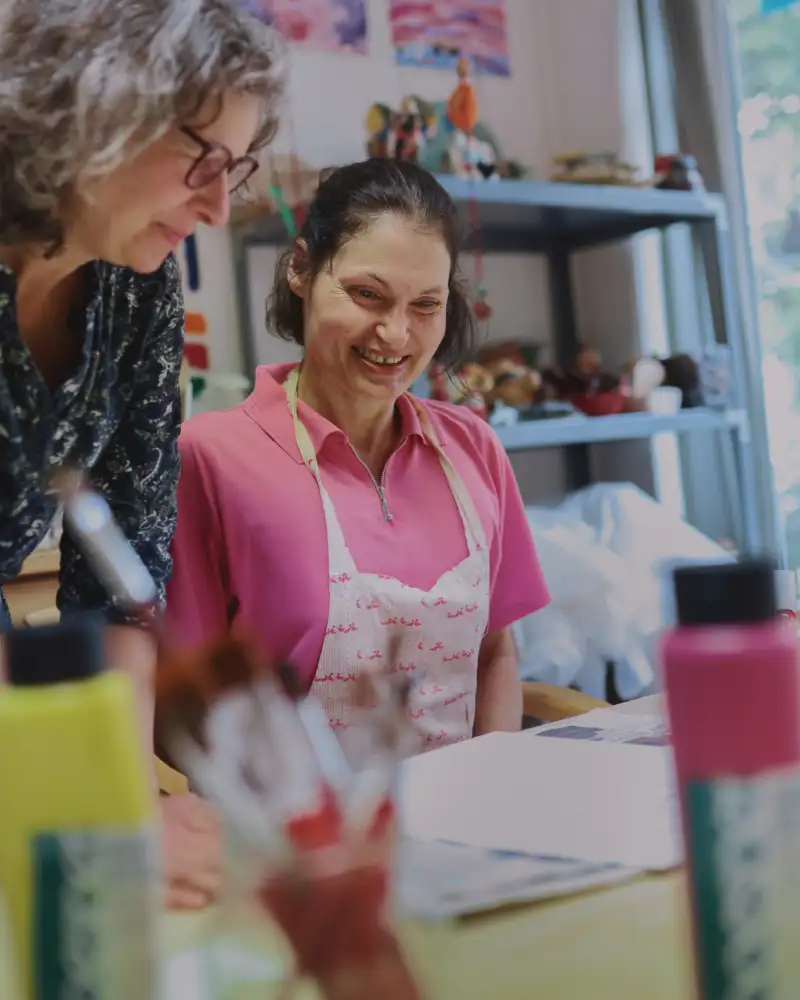Zwei Frauen sitzen an einem Tisch und schauen auf ein leeres Blatt Papier. Auf dem Tisch stehen Malutensilien.