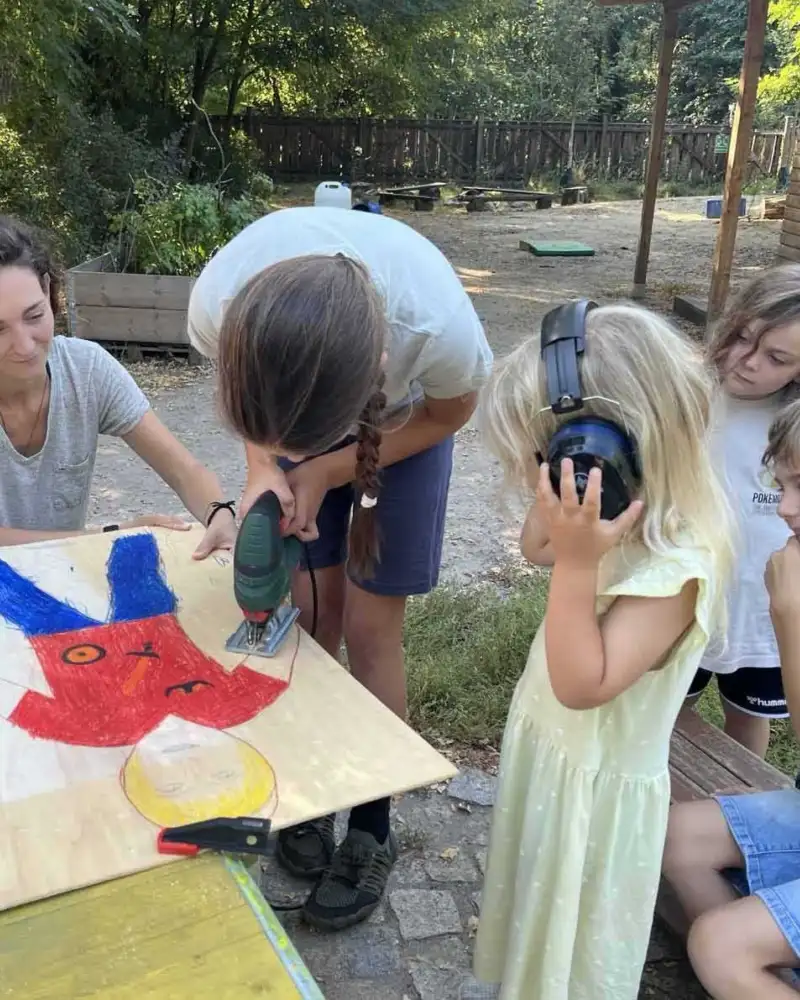 Eine Frau und mehrere Kinder arbeiten draußen an einem Tisch an einem Holzbild