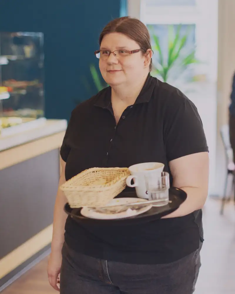 Eine Frau trägt ein Tablett mit Geschirr, im Hintergrund räumt ein Mann einen Tisch ab.