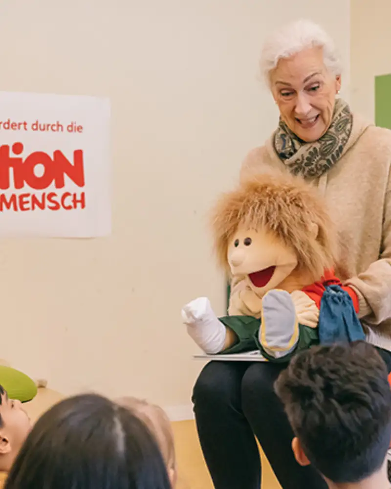 Zwei Frauen sitzen etwas erhöht vor einer Gruppe von Kindern. Jede von ihnen hat eine Handpuppe aus Stoff vor sich.