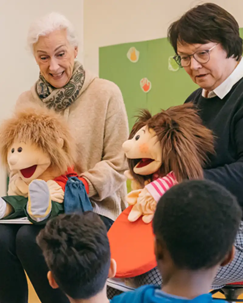 Zwei Frauen sitzen etwas erhöht vor einer Gruppe von Kindern. Jede von ihnen hat eine Handpuppe aus Stoff vor sich.