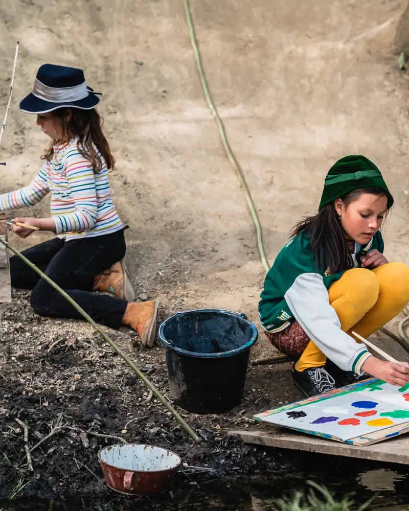 Zwei kleine Mädchen hocken an einem Hang und malen mit bunten Wasserfarben auf großen Papierbögen.